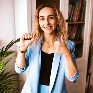 Kobieta o kręconych blond włosach ubrana w jasnoniebieski garnitur uśmiecha się do kamery, trzymając jedną pięść uniesioną w geście kciuka w górę, podobnym do symbolu w języku migowym. Stoi w pomieszczeniu w pobliżu rośliny i półki z książkami.