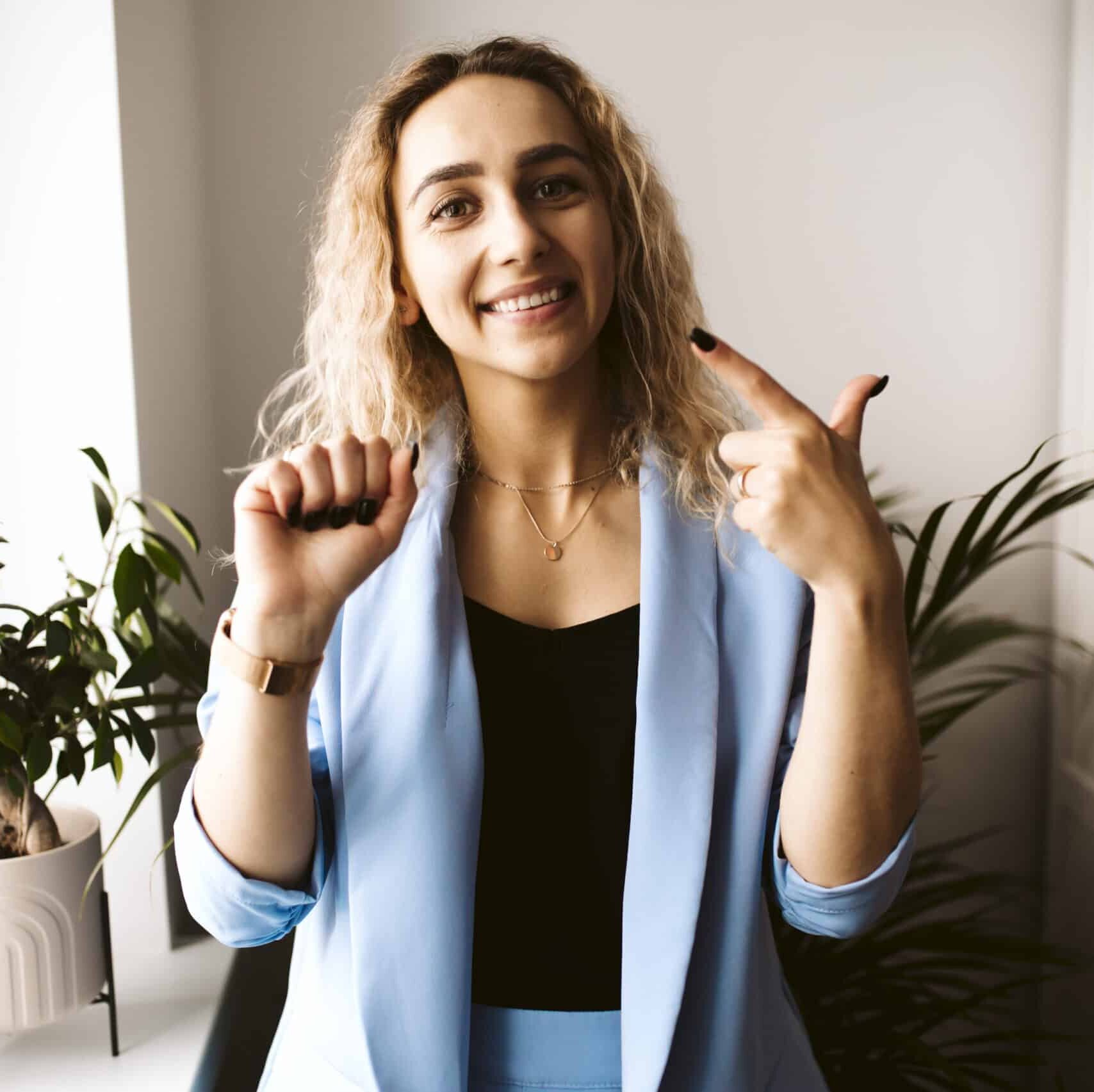 Uśmiechnięta kobieta z kręconymi blond włosami ubrana w jasnoniebieską marynarkę gestykuluje rękami, co wydaje się być językiem migowym. Stoi w pomieszczeniu w pobliżu roślin doniczkowych, oświetlona naturalnym światłem z pobliskiego okna.
