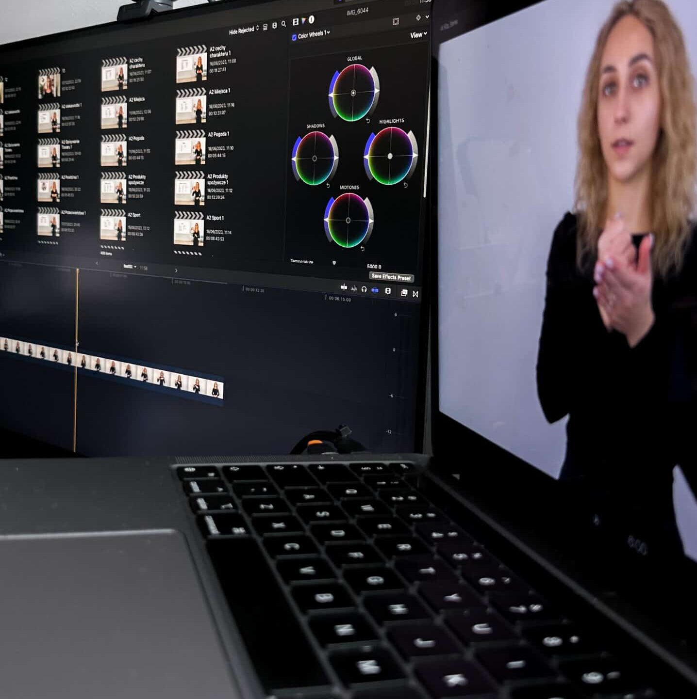 Zbliżenie laptopa pokazującego oprogramowanie do edycji wideo z kołami korekcji kolorów i klipami wideo, podczas gdy część ekranu przedstawia kobietę z kręconymi blond włosami w czarnej koszuli używającą języka migowego do gestykulacji rękami.